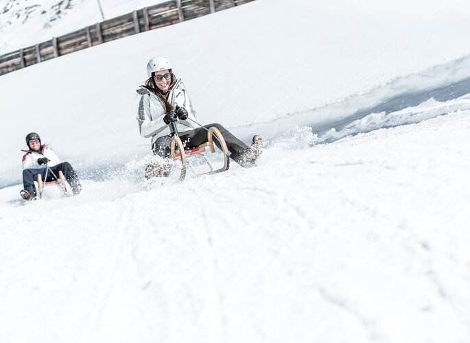 Zwei Personen rodeln auf einer verschneiten Winterbahn im Schnee.