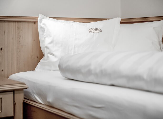 Ein weiß bezogenes Bett mit Hotel-Jenewein-Kissen neben einer Holzverkleidung.