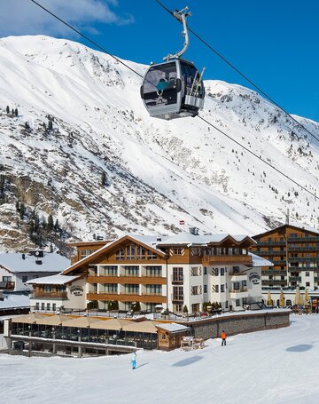 Das Hotel Jenewein liegt direkt an der Skipiste in Obergurgl unter einer Seilbahn vor verschneiten Bergen.