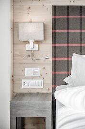 Ein Bett mit kariertem Kopfteil, Nachttisch und Wandleuchte in einem hellen Hotelzimmer.
