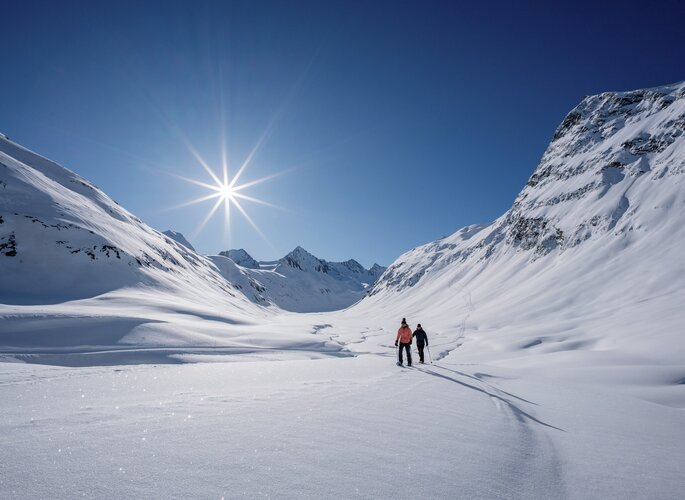 Zwei Personen wandern durch eine verschneite Winterlandschaft im Hochtal unter klarem Himmel.