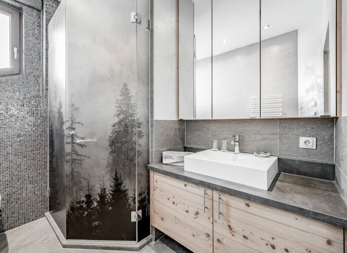 Komfortzimmer-Bad im Hotel Jenewein in Obergurgl Bad mit Waschbecken, Spiegel und Dusche im Komfortzimmer, gestaltet in hellen Grau- und Holztönen.
