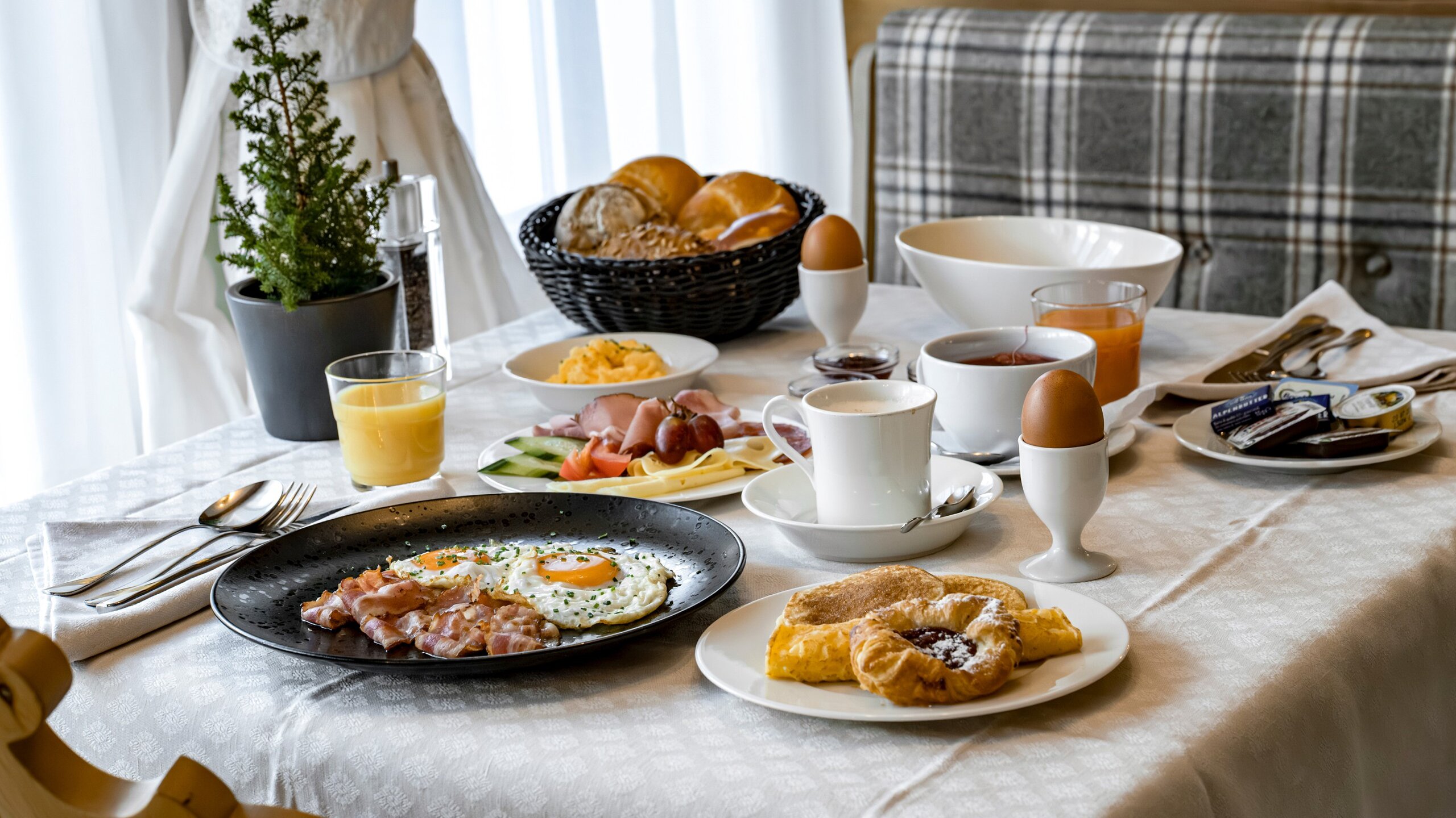 Ein gedeckter Frühstückstisch mit Eiern, Speck, Gebäck, Brot, Aufschnitt, Saft und Kaffee.