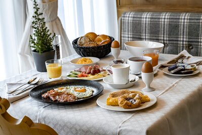 Ein reich gedeckter Frühstückstisch mit Eiern, Gebäck, Aufschnitt, Saft und Kaffee am Fenster.