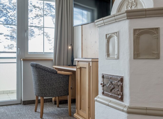 Appartement mit Kachelofen im Landhaus Jenewein Ein Schreibtisch mit Stuhl steht neben dem Kachelofen und dem Fenster mit Blick auf die verschneite Berglandschaft.