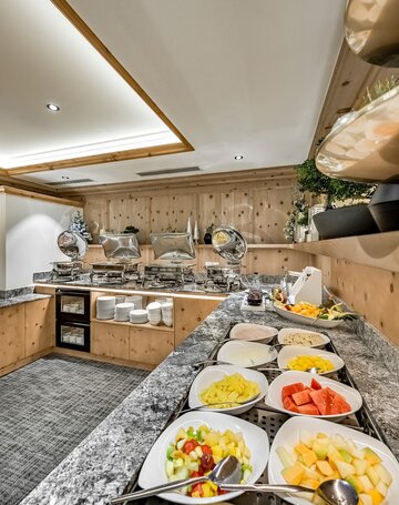Ein Frühstücksbuffet mit Obst, Joghurt und Warmhaltebehältern in einem hellen Speiseraum.