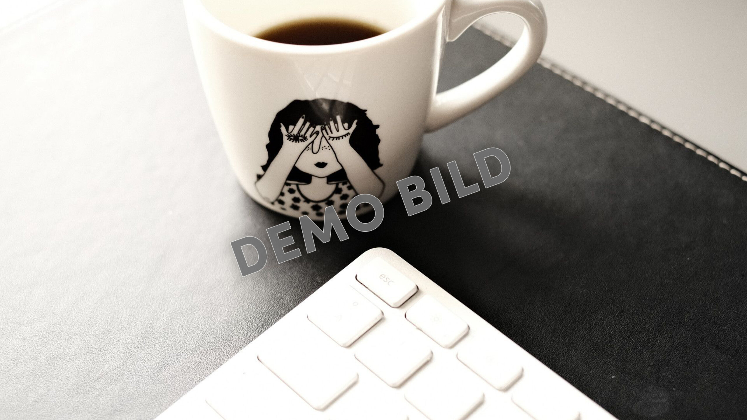 Eine weiße Kaffeetasse steht neben einer weißen Tastatur auf einer dunklen Schreibtischunterlage.