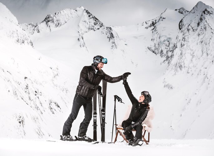 Zwei Skifahrer genießen eine Pause im Schnee vor einer verschneiten Bergkulisse.