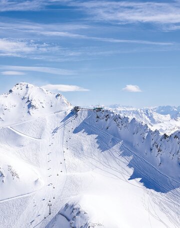 Schneebedeckte Skipisten und ein Sessellift auf einem Berggipfel unter blauem Himmel.