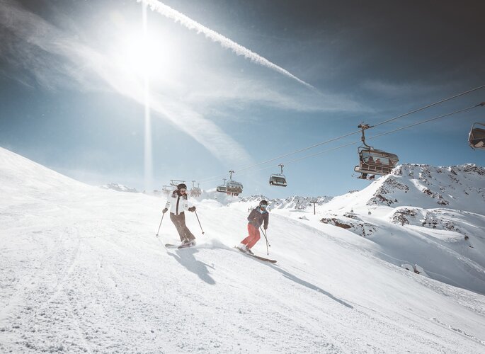 Zwei Skifahrer fahren unter einem Sessellift durch eine verschneite Piste im sonnigen Hochgebirge.