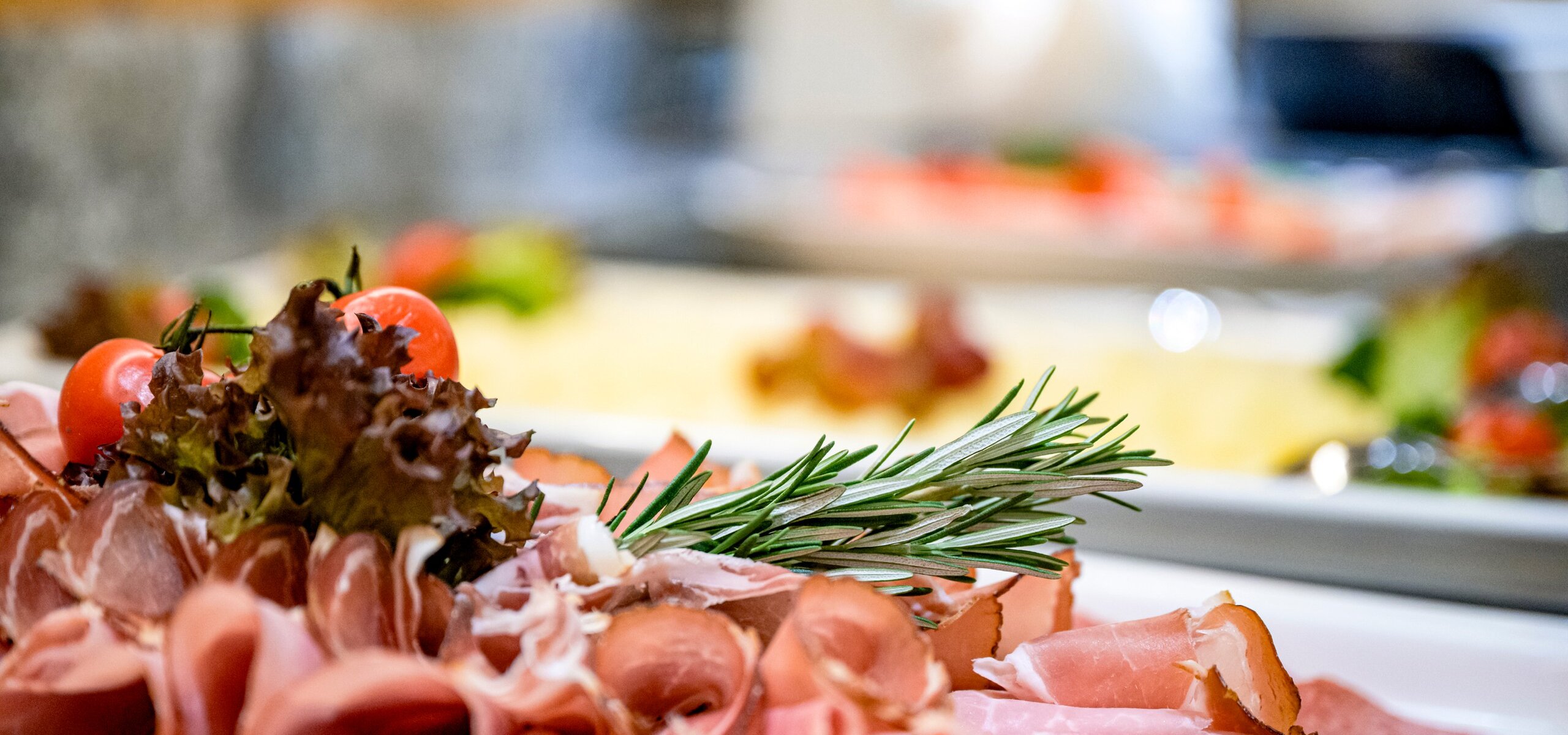 Ein Frühstücksbuffet mit Aufschnitt, Tomaten, Salat und frischen Kräutern ist dekorativ angerichtet.