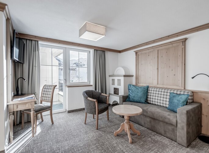 Heller Wohnbereich im Familienappartement mit Sitzgruppe, Kachelofen und Blick auf die verschneiten Berge.