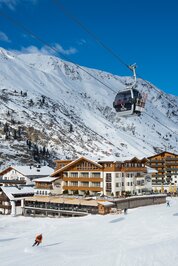 Eine Seilbahnkabine schwebt über dem Hotel Jenewein im verschneiten Obergurgl, davor fährt ein Skifahrer talwärts.