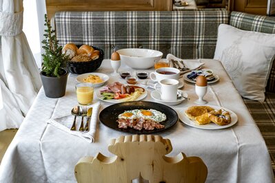 Ein reich gedeckter Frühstückstisch mit Eiern, Gebäck, Aufschnitt, Saft und Kaffee am Fenster.