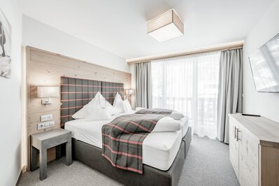 Helles Hotelzimmer mit Doppelbett, Holzverkleidung und großen Fenstern im Hotel Jenewein in Obergurgl.