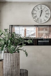Ein Pflanzenkübel steht vor einer Wand mit Uhr und Spiegel im Ruhebereich.
