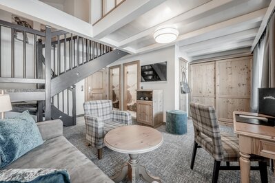 Ein heller Wohnbereich einer Suite mit Sofa, Sesseln, Holzmöbeln und Treppe im Hotel Jenewein.