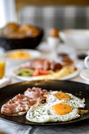 Ein Teller mit Speck und Spiegeleiern im Frühstücksbereich, im Hintergrund weitere Frühstückszutaten.