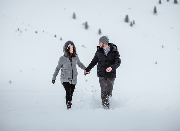 Ein Paar wandert durch tiefen Schnee in einer ruhigen Winterlandschaft mit einzelnen Bäumen im Hintergrund.