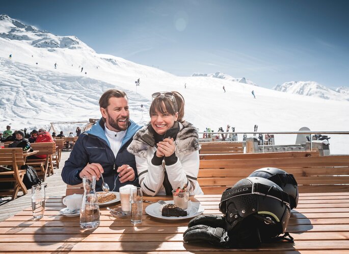 Skiurlaub mit Hüttengaudi in Obergurgl Ein Paar sitzt auf einer Sonnenterrasse im Skigebiet und genießt eine Pause mit Blick auf die verschneiten Hänge.