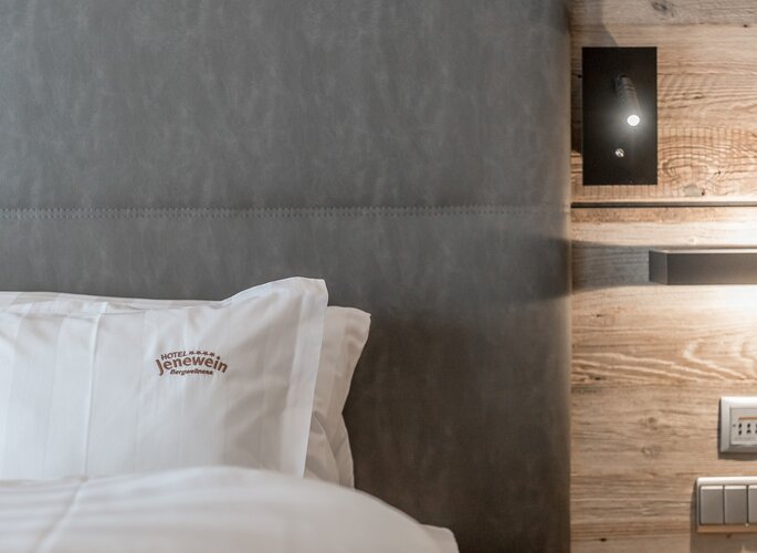 Romantikzimmer im Hotel Jenewein in Obergurgl Ein Bett mit Kissen im Romantikzimmer des Hotel Jenewein, daneben eine Holzverkleidung und eine dezente Wandlampe.