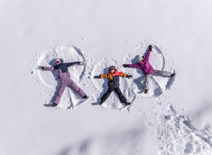 Drei Personen liegen im frischen Schnee und formen Schneekreise mit ausgebreiteten Armen.