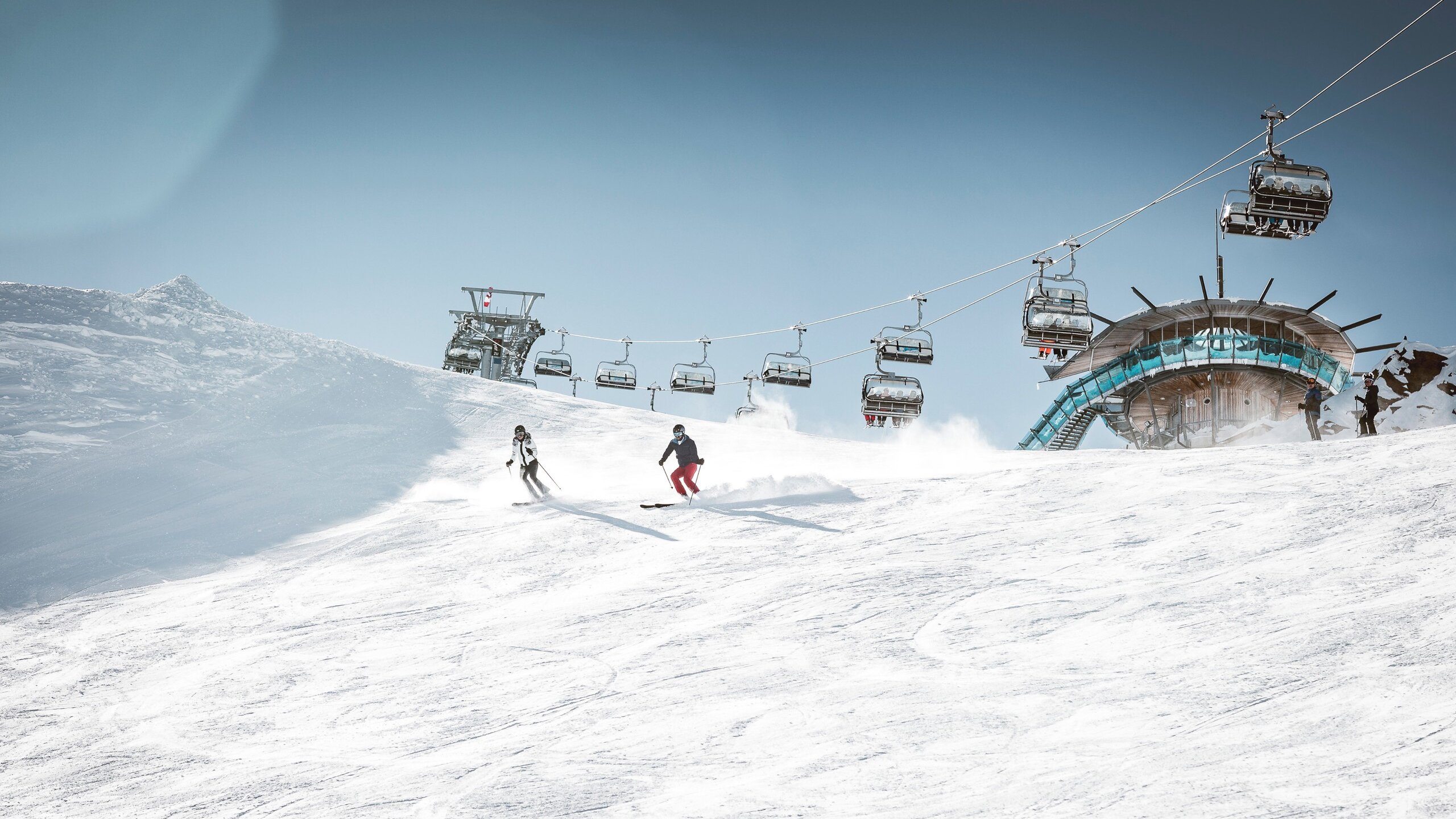 Zwei Skifahrer fahren auf einer verschneiten Piste unter der Bergbahn im Skigebiet Gurgl.