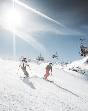 Zwei Skifahrer fahren auf einer sonnigen Skipiste unter einem Sessellift ab.