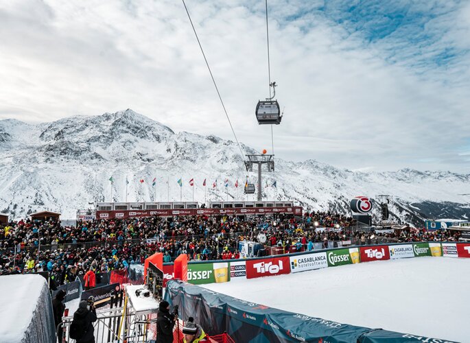 Eine große Menschenmenge verfolgt eine Winterveranstaltung im Schnee, während eine Seilbahn über die Tribüne fährt.
